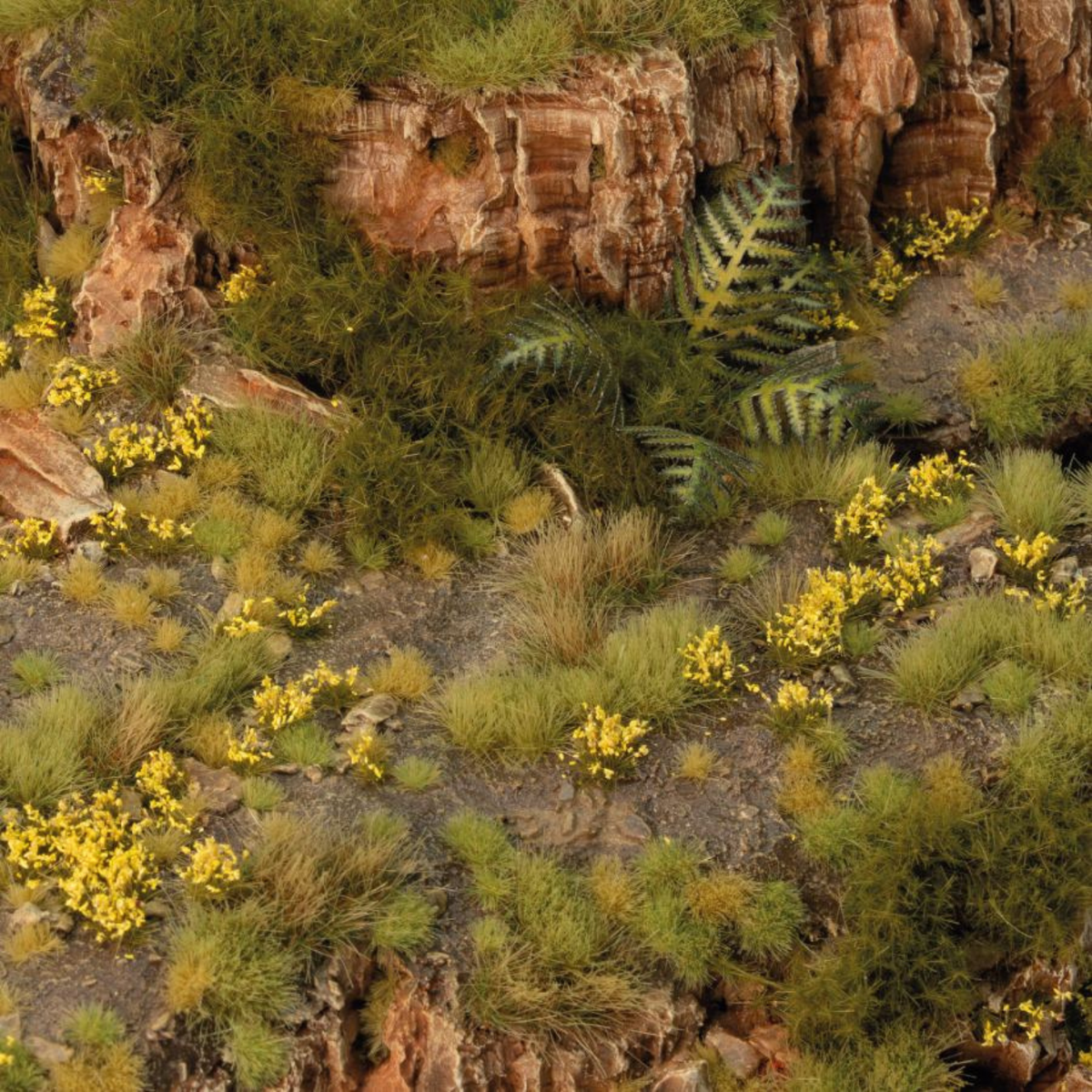 A close-up of a textured tabletop terrain or diorama scene featuring rocky cliffs, green grasses, and small clusters of yellow wildflowers.