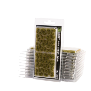 Packaged dry green grass miniature foam pieces in a clear blister pack with a white cardboard header, shown with stacks of the same product in the background on a white studio background.