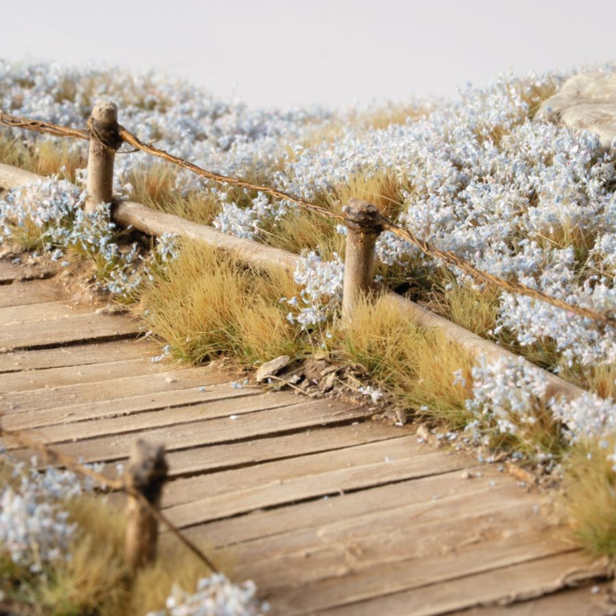 A miniature diorama scale model of a wooden boardwalk path with a rustic wooden fence and wire railing, surrounded by detailed grass and white flowers.