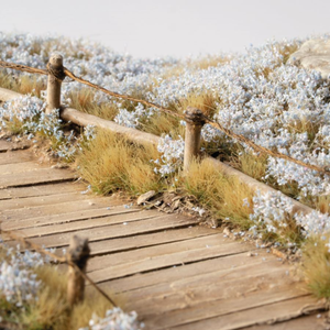 A miniature diorama scale model of a wooden boardwalk path with a rustic wooden fence and wire railing, surrounded by detailed grass and white flowers.