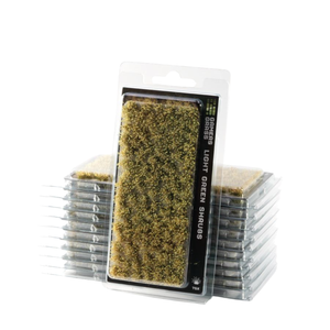 Packaged miniature light green shrubs for tabletop wargaming, shown in clear plastic blister packs with a black label, with additional unopened packs stacked behind.