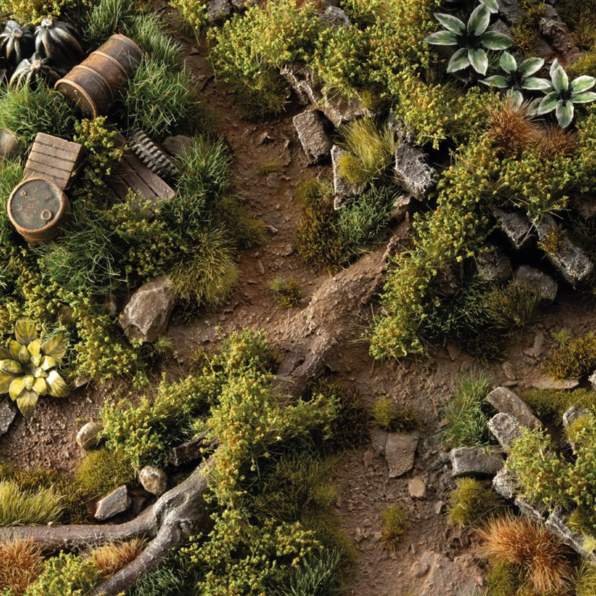 A detailed miniature terrain scene with a dirt path winding through dense green moss, grass, rocks, and small decorative props like barrels and wooden crates.