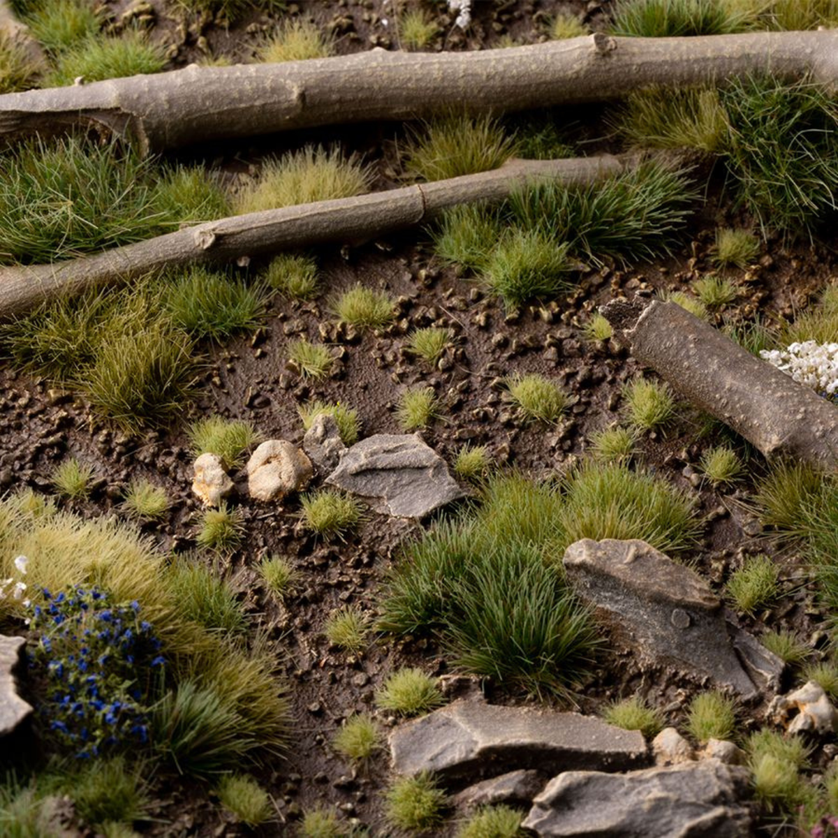 Naturalistic miniature terrain featuring green grass tufts, brown soil, scattered gray rocks, and two fallen logs in a forest-like setting.