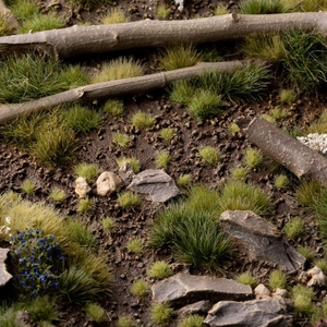 Naturalistic miniature terrain featuring green grass tufts, brown soil, scattered gray rocks, and two fallen logs in a forest-like setting.