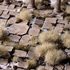 A close-up of a miniature terrain mat featuring detailed stone paving stones and tufts of dry grass, with a classical column base visible in the background.